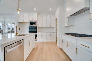 Kitchen with stainless steel appliances, custom exhaust hood, light wood-style floors, light stone counters, and decorative light fixtures