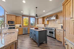 Kitchen featuring dark wood-style flooring, stainless steel appliances, wall chimney exhaust hood, recessed lighting, and pendant lighting