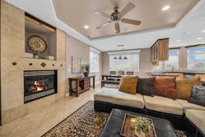 Living area with a tiled fireplace, light tile patterned floors, a ceiling fan, a tray ceiling, and recessed lighting