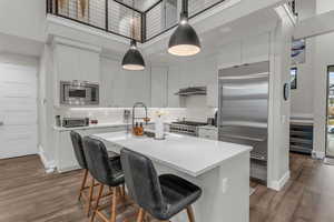 Kitchen featuring a breakfast bar area, built in appliances, a kitchen island with sink, pendant lighting, and white cabinetry