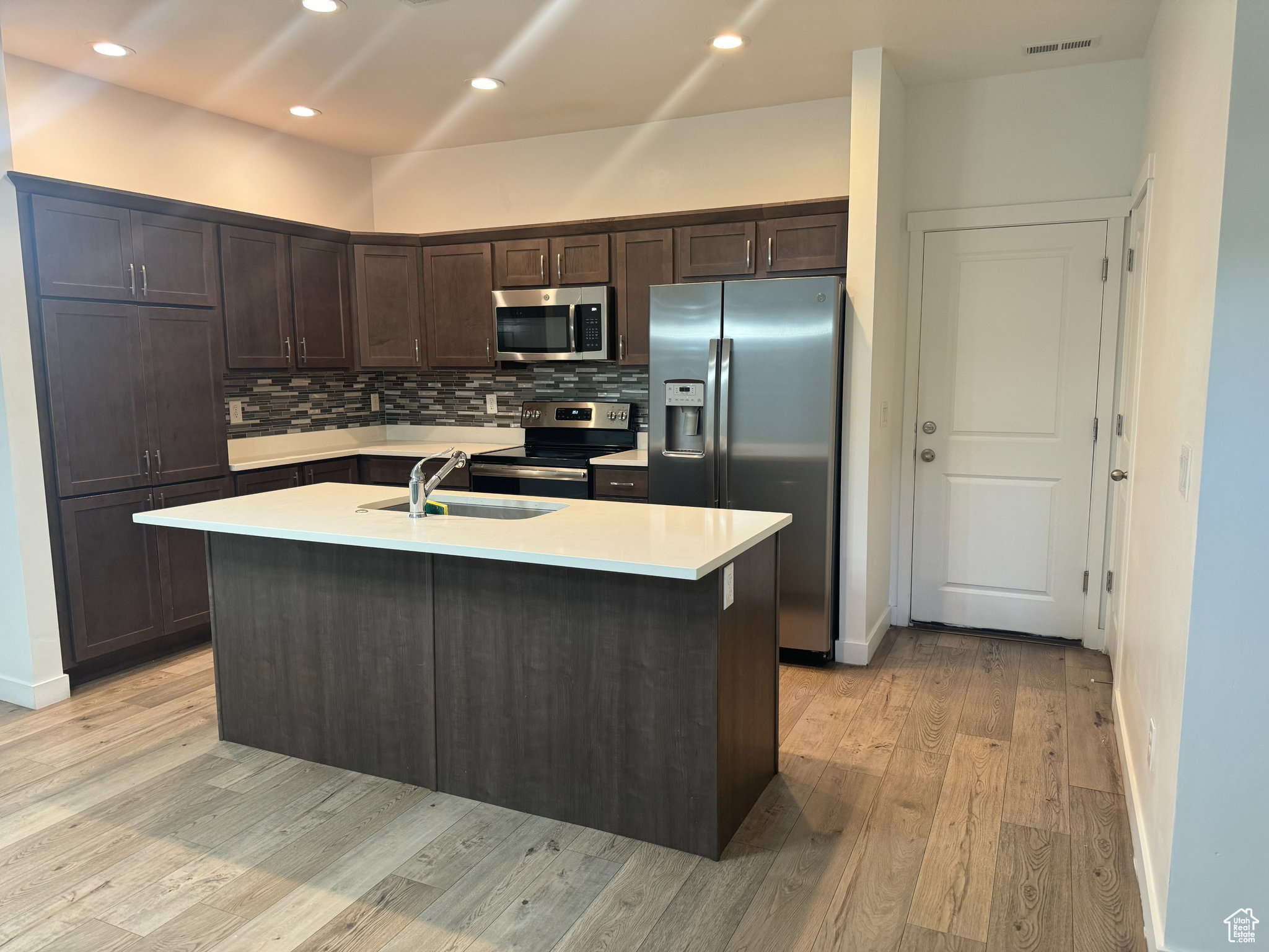 Kitchen with dark brown cabinetry, stainless steel appliances, tasteful backsplash, a center island with sink, and recessed lighting