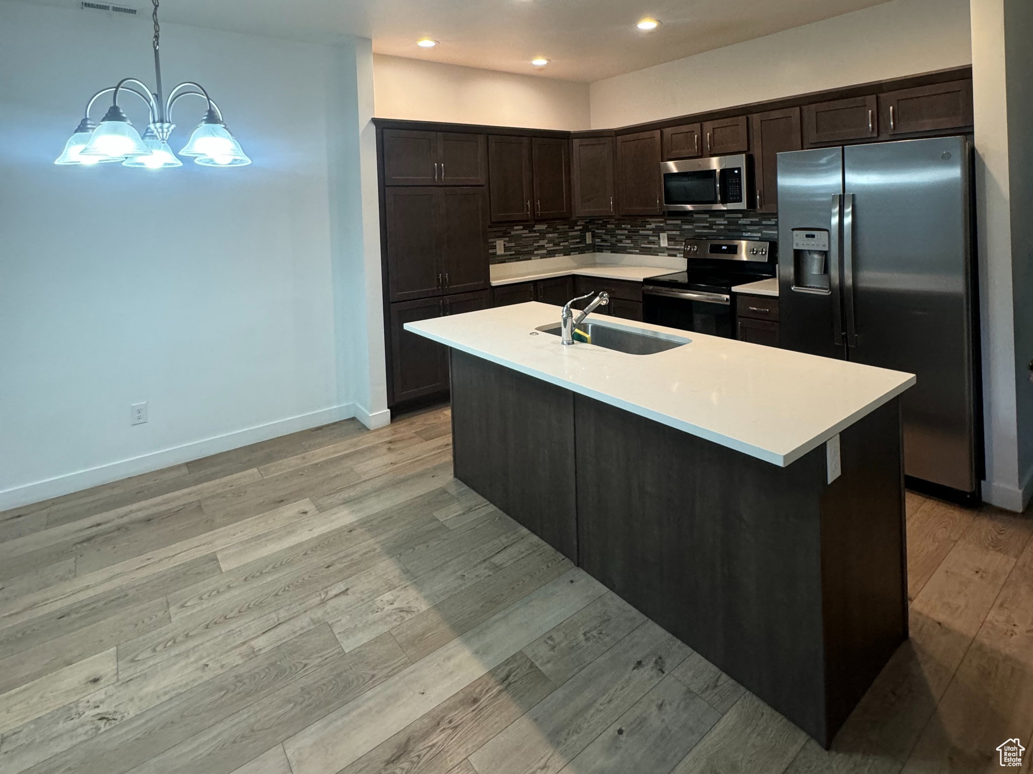 Kitchen with appliances with stainless steel finishes, dark brown cabinets, backsplash, hanging light fixtures, and a kitchen island with sink