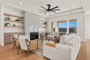 Living area featuring built in features, a premium fireplace, light wood-style flooring, a raised ceiling, and a ceiling fan