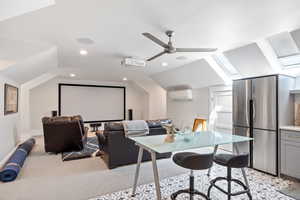Home theater room featuring vaulted ceiling, a skylight, a wall mounted air conditioner, light colored carpet, and ceiling fan