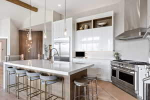 Kitchen with wall chimney exhaust hood, premium appliances, hanging light fixtures, white cabinets, and an island with sink