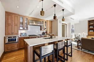 Kitchen with a kitchen breakfast bar, decorative light fixtures, glass insert cabinets, wall chimney range hood, and a kitchen island with sink