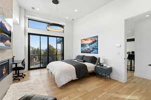 Bedroom with access to exterior, light wood-style flooring, a high ceiling, recessed lighting, and a glass covered fireplace
