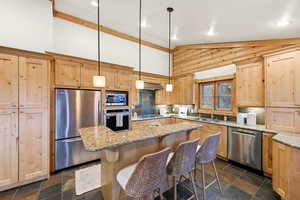 Kitchen featuring light brown cabinetry, light stone counters, stainless steel appliances, decorative light fixtures, and a kitchen breakfast bar
