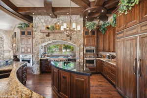 Kitchen with dark stone countertops, beam ceiling, glass insert cabinets, paneled built in refrigerator, and dark wood-style flooring