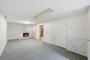 Unfurnished living room featuring a fireplace, a textured ceiling, carpet flooring, a wainscoted wall, and brick wall