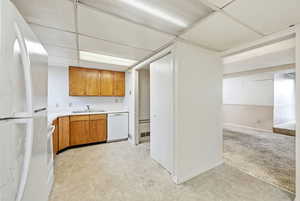 Kitchen featuring white appliances, light countertops, brown cabinetry, light colored carpet, and a drop ceiling