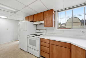 Kitchen featuring white appliances, light countertops, brown cabinetry, under cabinet range hood, and a drop ceiling