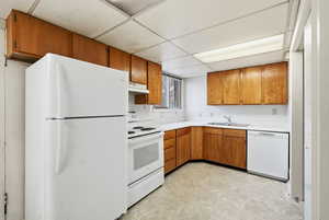 Kitchen featuring brown cabinetry, white appliances, light countertops, a drop ceiling, and light flooring