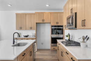Kitchen with light wood finish cabinetry, light stone countertops, stainless steel appliances, light wood-type flooring, and a center island with sink