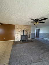 Unfurnished living room with a wood stove, ceiling fan, tile patterned flooring, a wainscoted wall, and a textured ceiling