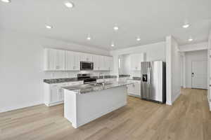 Kitchen featuring white cabinets, appliances with stainless steel finishes, dark stone countertops, an island with sink, and light wood-type flooring