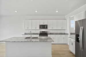 Kitchen with stainless steel appliances, light stone counters, white cabinets, a center island with sink, and recessed lighting