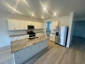 Kitchen with appliances with stainless steel finishes, white cabinets, light stone counters, an island with sink, and light wood-style flooring