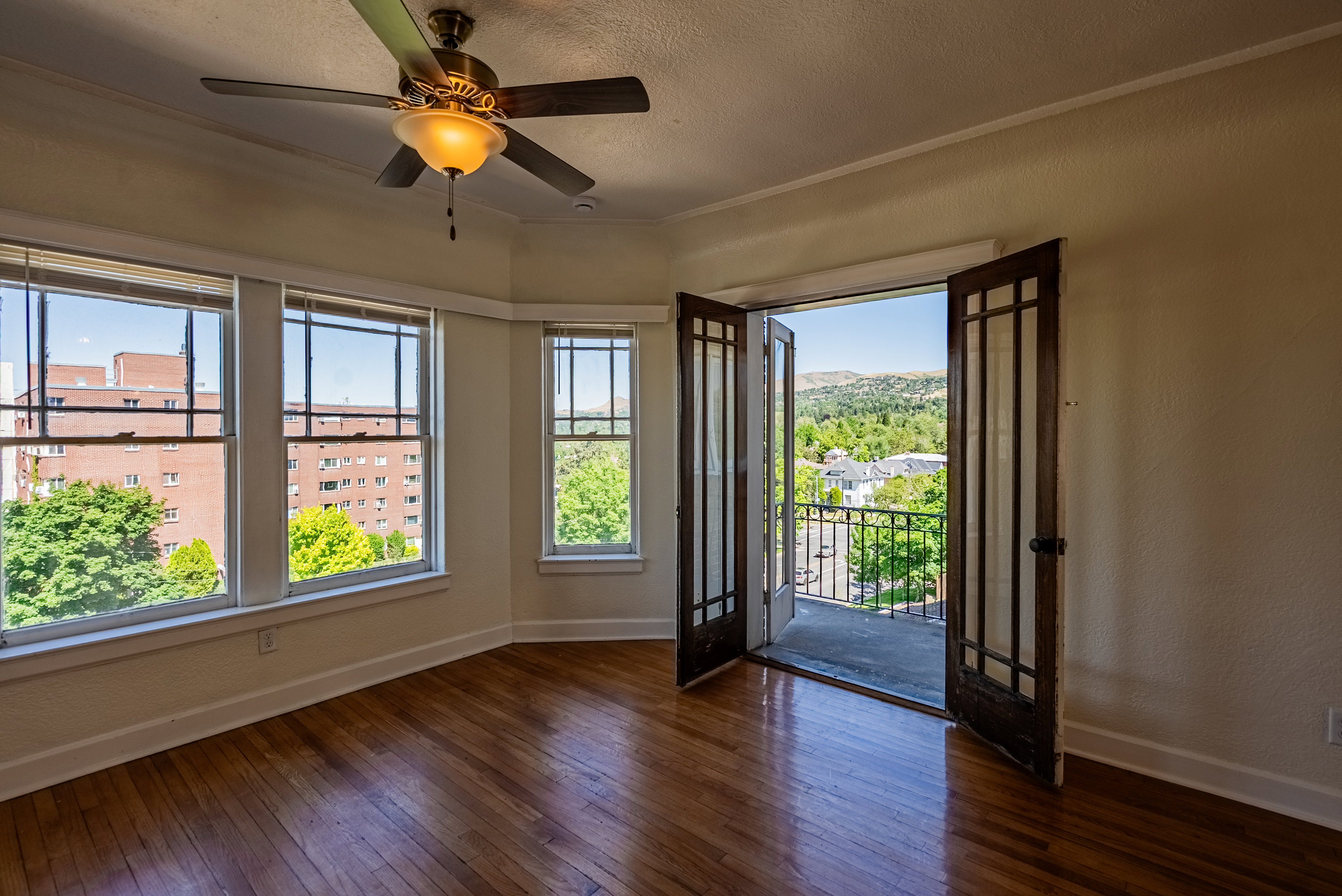 Living space featuring wood-finish flooring, a ceiling fan with integrated lighting, multiple windows, and exterior access via double doors to a balcony with metal railing
