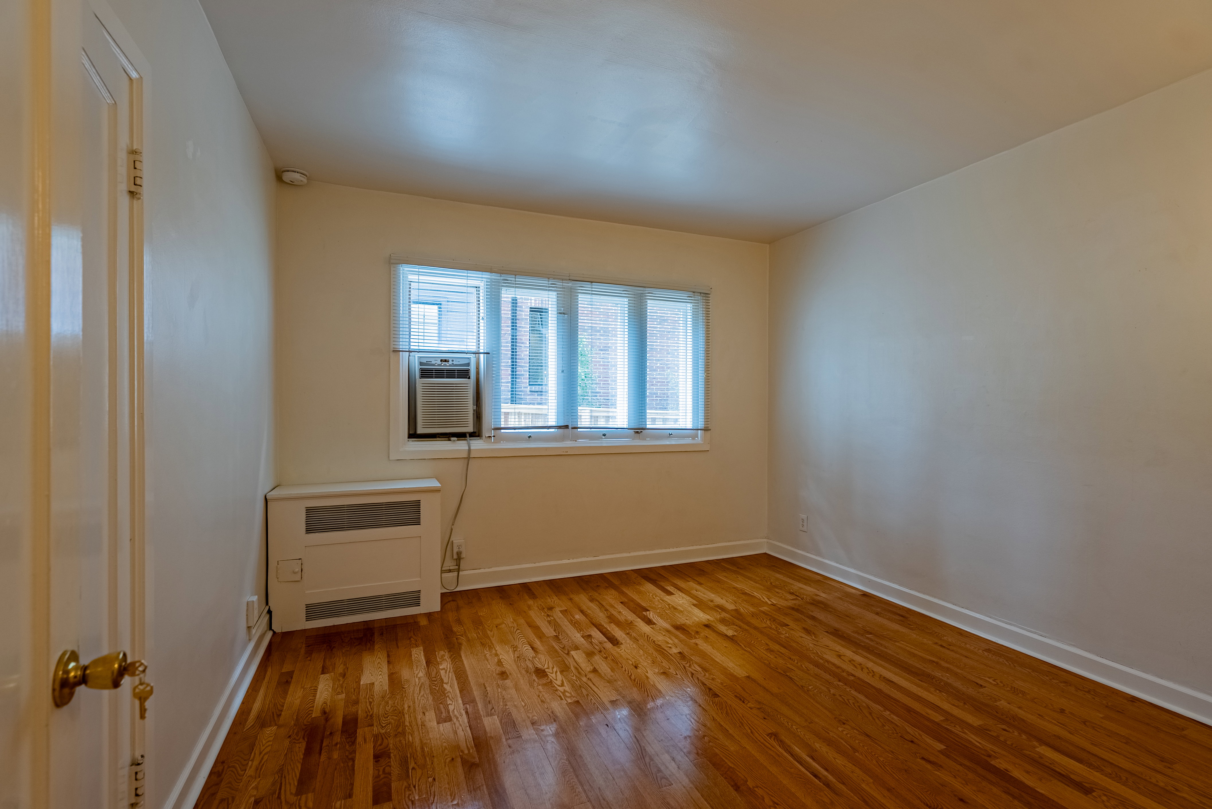 Hardwood flooring in a neutral-toned room featuring a window with blinds, a window-mounted air conditioning unit, and a built-in heating unit