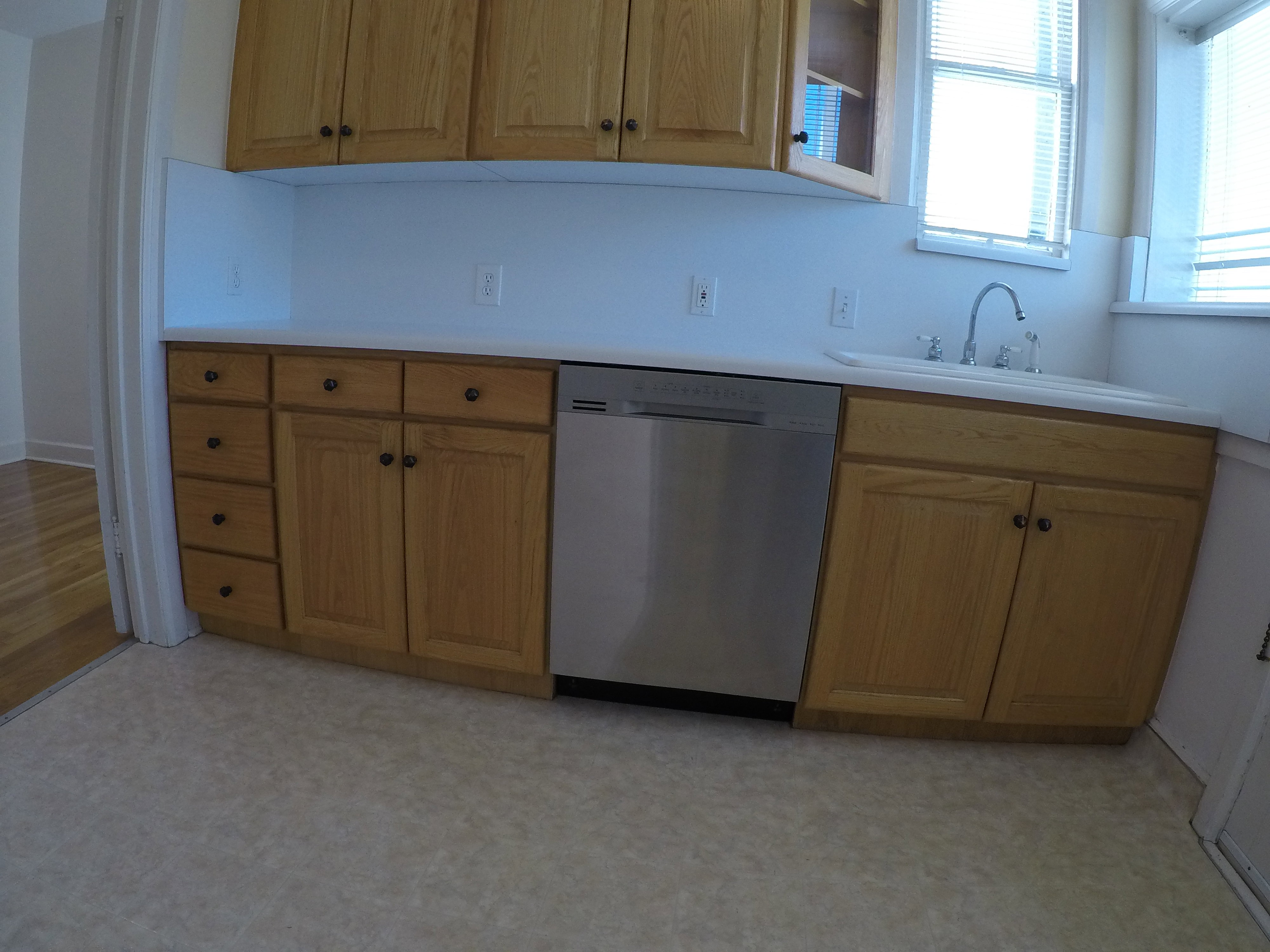 Apt # 8 > Kitchen featuring stainless steel dishwasher, light countertops, glass insert cabinets, light flooring, and brown cabinets