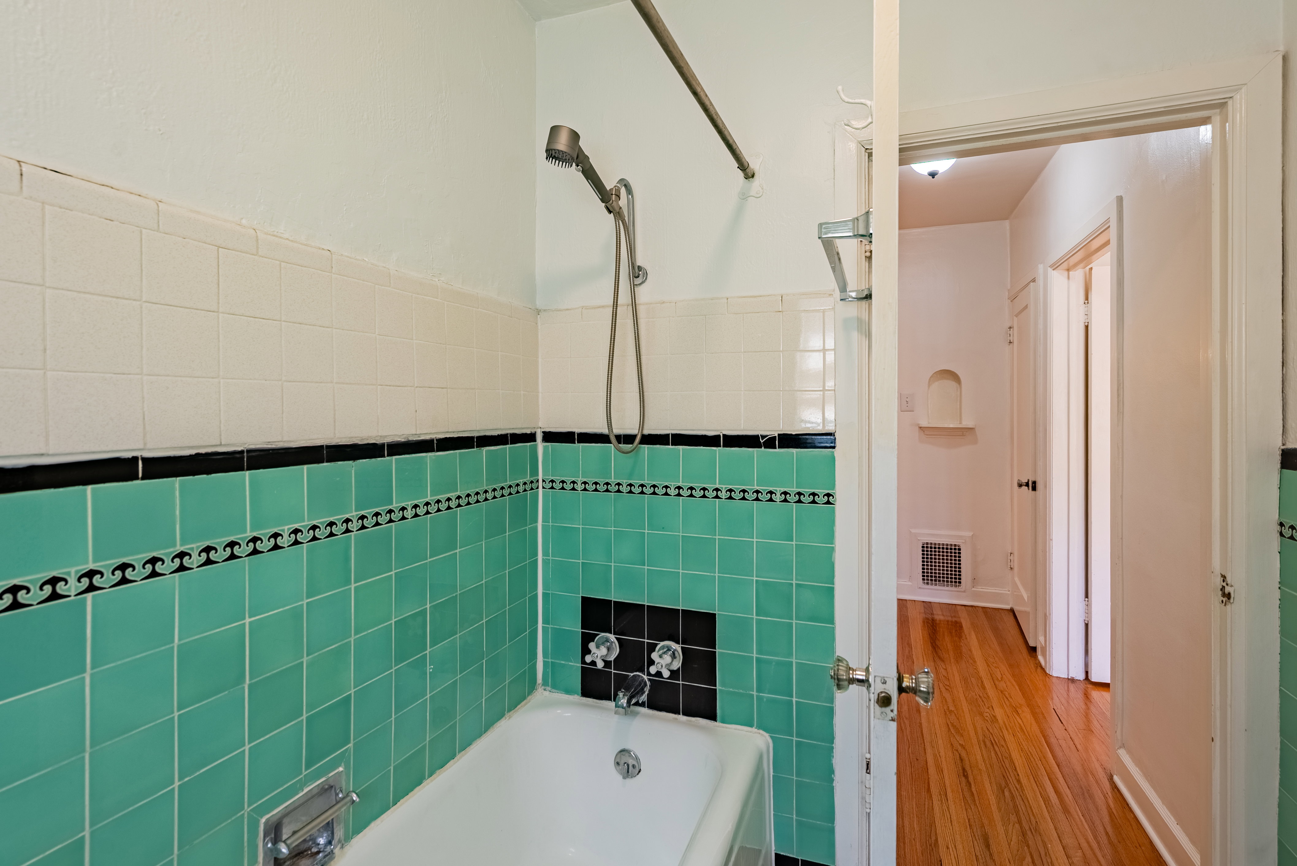 Bathroom featuring a white bathtub with chrome fixtures, a tiled shower surround with teal and black accent tiles, a handheld showerhead, and white walls