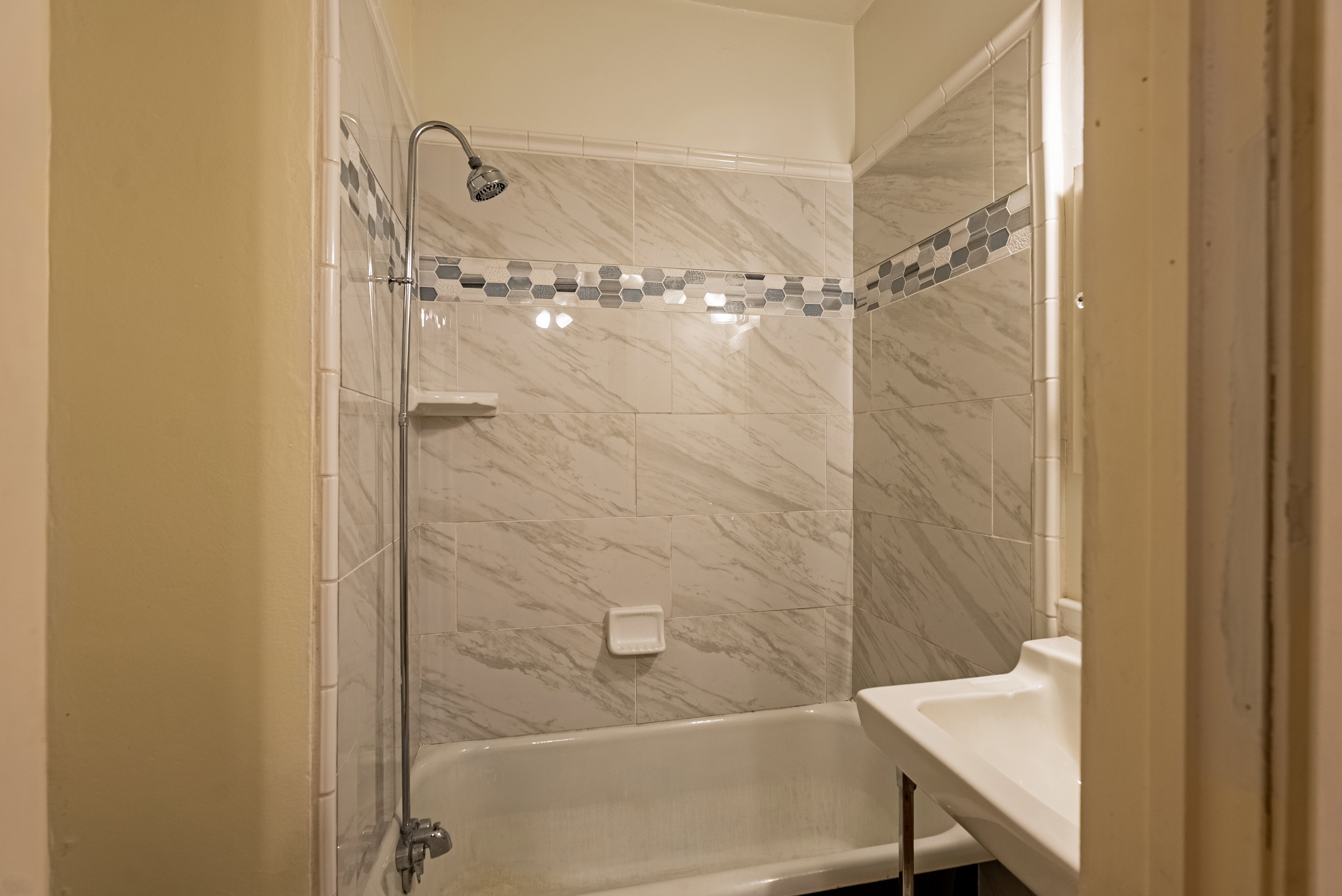 Bathroom featuring a built-in bathtub with a shower head, white subway tile surround, decorative mosaic tile accent, and a pedestal sink