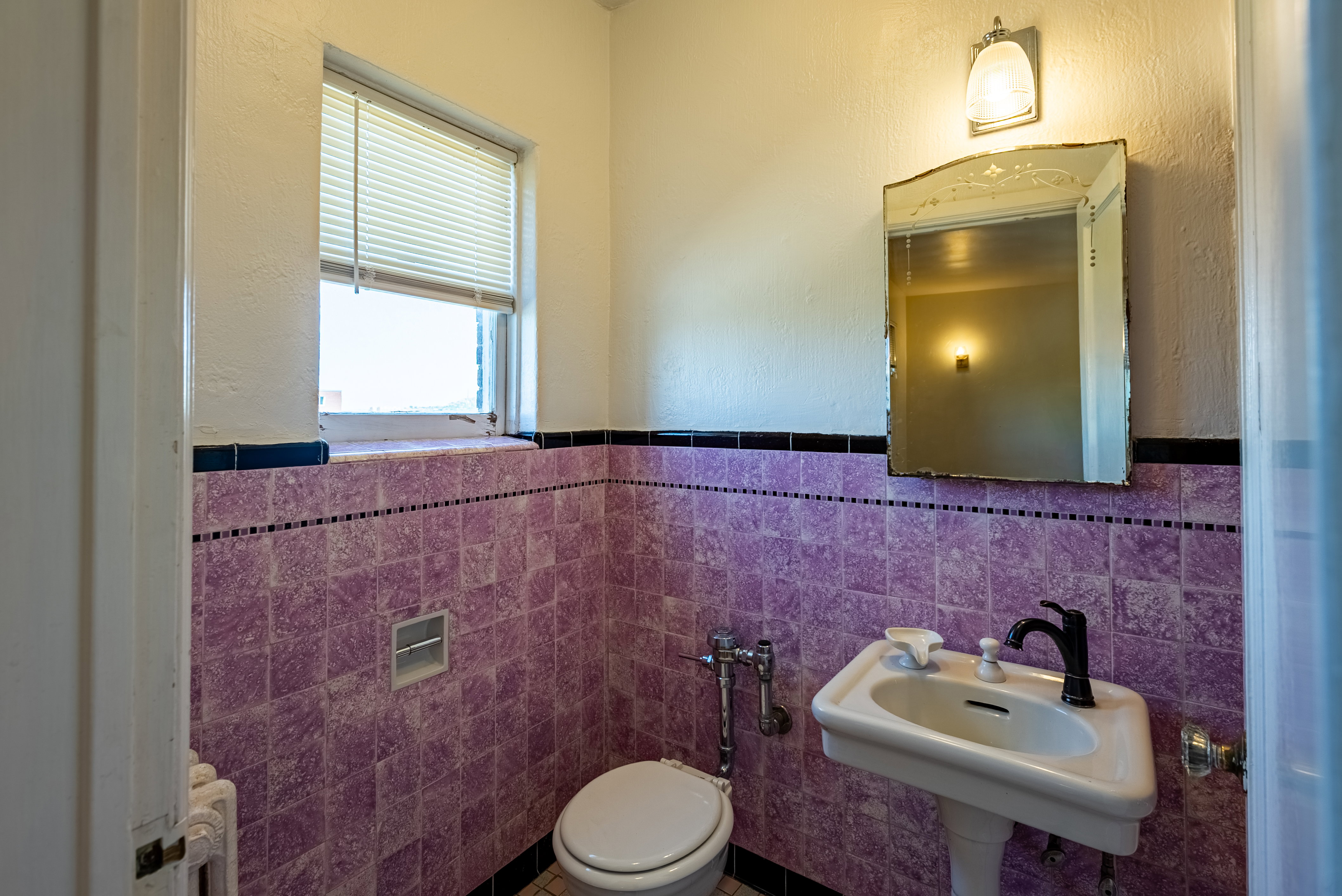 Bathroom featuring full-height purple wall tile with black trim, a pedestal sink with a black faucet, and a wall-mounted mirror with an overhead light fixture