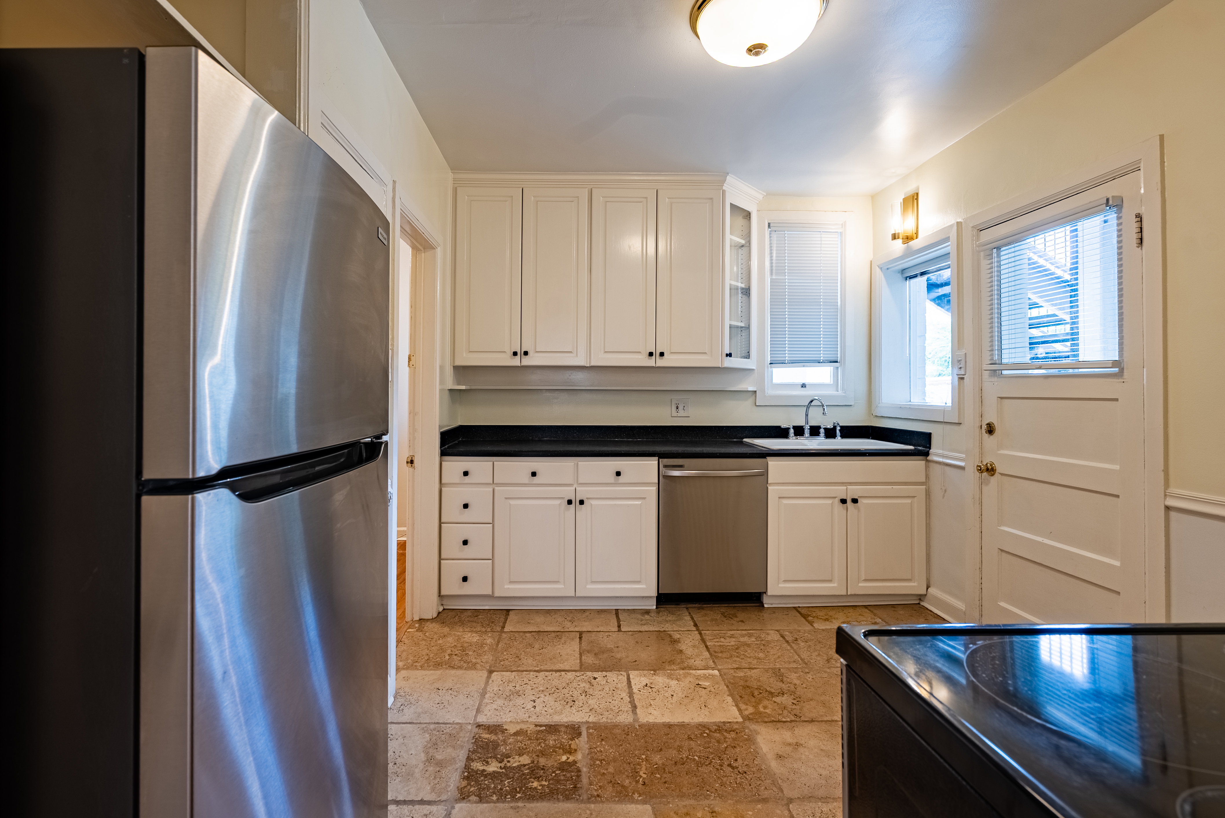 Kitchen featuring white cabinetry, dark countertops, a stainless steel dishwasher, and stone tile flooring