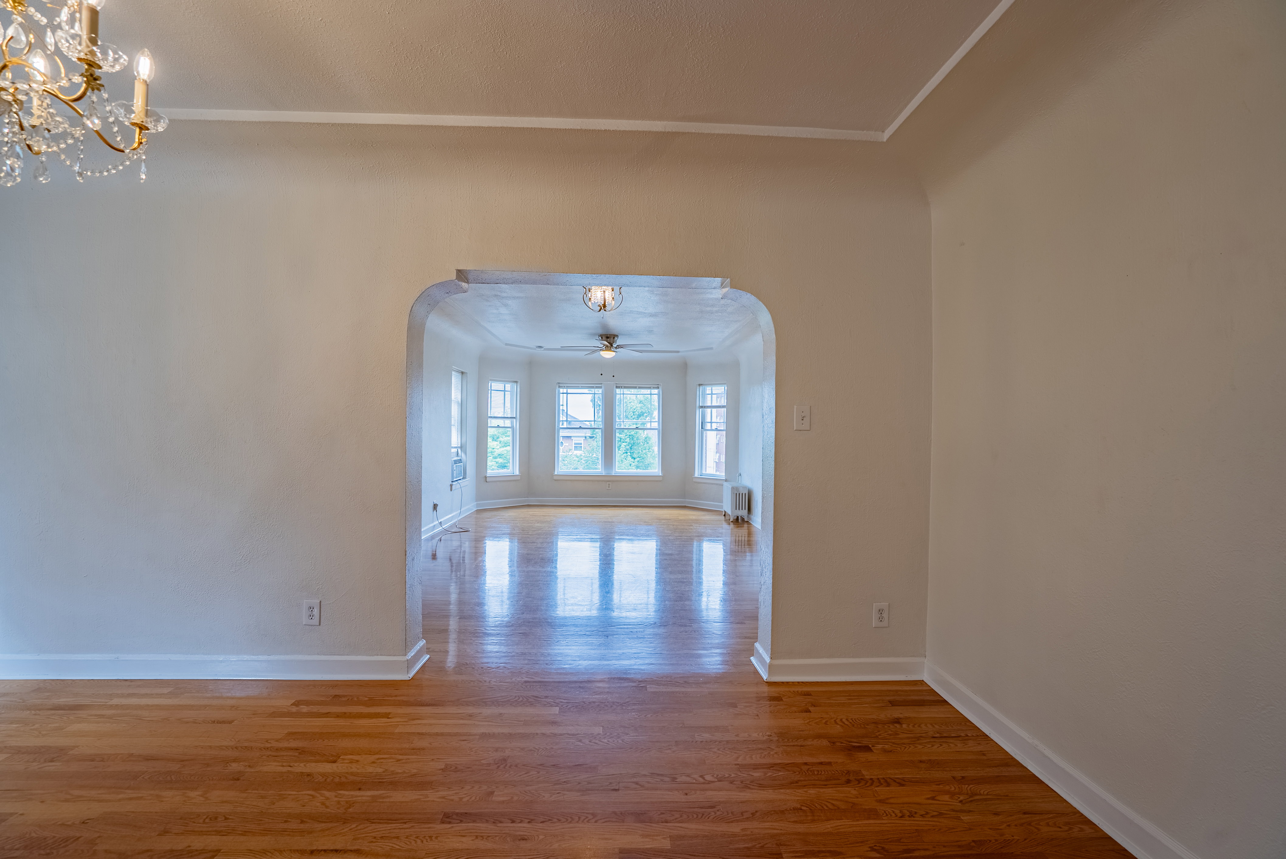 Wood-finish flooring throughout, featuring a chandelier with crystal accents and an arched entryway leading to a sunlit bay window area with multiple windows and a ceiling fan