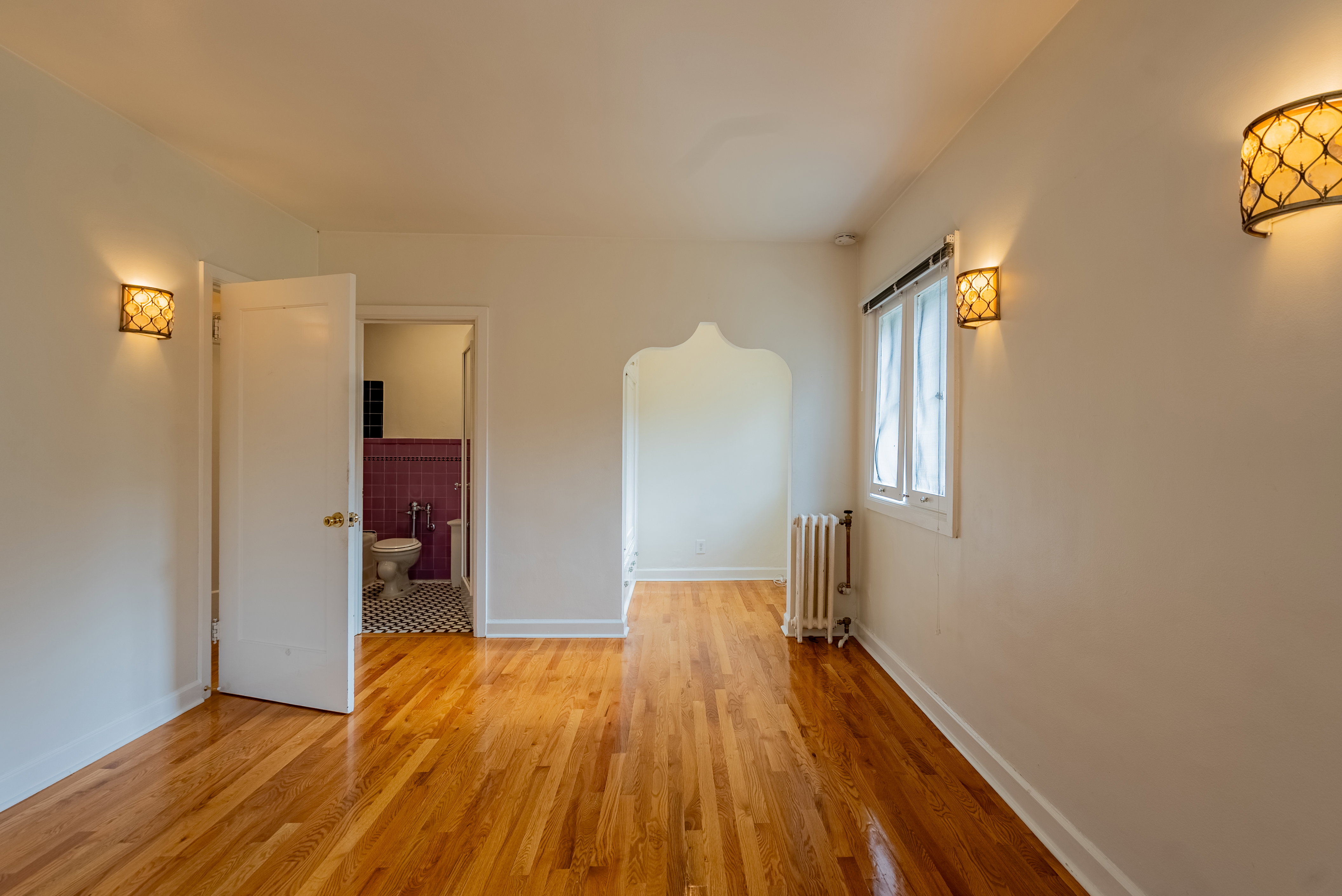 Hardwood flooring throughout featuring sconce lighting, arched doorway, and window with radiator