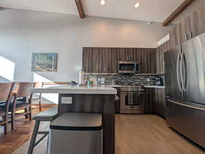 Kitchen with stainless steel appliances, beamed ceiling, a kitchen bar, light countertops, and dark brown cabinetry