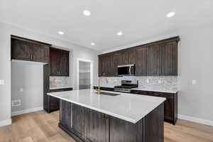 Kitchen featuring tasteful backsplash, dark brown cabinets, light stone counters, stainless steel appliances, and an island with sink