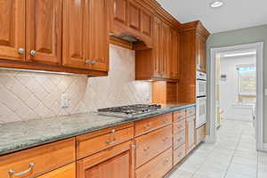 Kitchen featuring brown cabinetry, dark stone countertops, light tile patterned floors, tasteful backsplash, and double oven