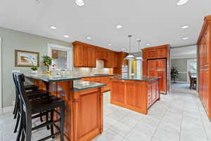 Kitchen with dark stone counters, brown cabinetry, decorative light fixtures, a kitchen island, and a kitchen breakfast bar
