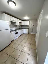 Kitchen featuring white appliances, white cabinetry, light tile patterned floors, under cabinet range hood, and dark countertops