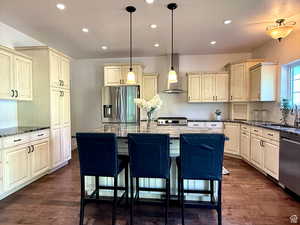 Kitchen featuring cream cabinetry, dark stone counters, stainless steel appliances, wall chimney range hood, and recessed lighting