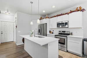 Kitchen featuring stainless steel appliances, white cabinetry, decorative light fixtures, a kitchen island with sink, and backsplash