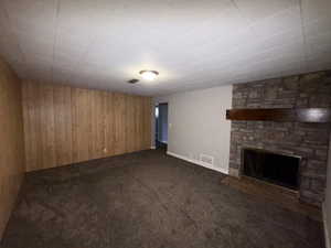 Unfurnished living room featuring wood walls, a stone fireplace, and dark colored carpet