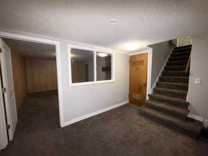 Finished basement with a textured ceiling, dark colored carpet, stairs, and wood walls