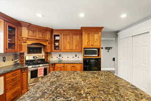 Kitchen with brown cabinetry, glass insert cabinets, stainless steel appliances, dark stone counters, and recessed lighting