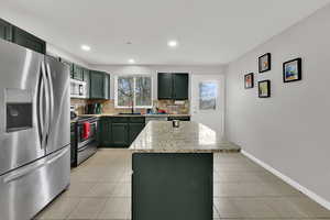 Kitchen with appliances with stainless steel finishes, green cabinets, tasteful backsplash, a center island, and light stone counters