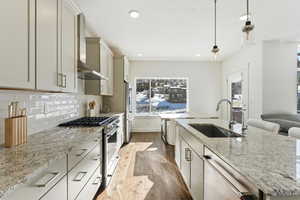 Kitchen with light stone counters, appliances with stainless steel finishes, dark wood-type flooring, pendant lighting, and wall chimney range hood
