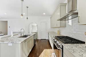 Kitchen featuring stainless steel appliances, wall chimney range hood, light stone countertops, arched walkways, and hanging light fixtures