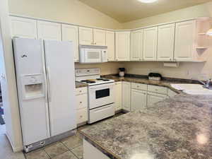 Kitchen featuring dark countertops, white appliances, lofted ceiling, white cabinets, and light tile patterned floors