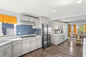 Kitchen featuring appliances with stainless steel finishes, gray cabinetry, dark wood finished floors, under cabinet range hood, and tasteful backsplash