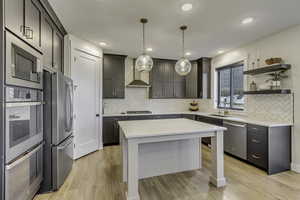 Kitchen with hanging light fixtures, appliances with stainless steel finishes, gray cabinets, light wood finished floors, and wall chimney range hood