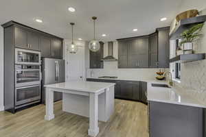Kitchen featuring hanging light fixtures, stainless steel appliances, wall chimney exhaust hood, light stone countertops, and a center island