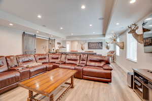 Living room with a barn door, a raised ceiling, light wood finished floors, recessed lighting, and healthy amount of natural light