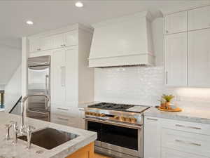 Kitchen with premium appliances, custom exhaust hood, white cabinets, light stone countertops, and tasteful backsplash